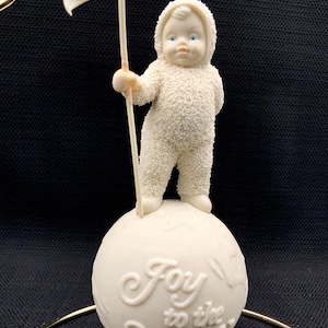 May include: A white textured figurine of a child in a hooded snowsuit standing on a spherical base. The child holds a flag with star details. The base features the embossed text Joy to the World. The figurine has blue eyes and a smooth finish.