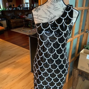 May include: A black and white one-shoulder sequined dress with a mermaid-style skirt. The sequins are arranged in a scale pattern.