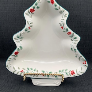 Puede incluir: Plato de servir de cerámica blanca con forma de árbol de Navidad con acebo verde y bayas rojas alrededor del borde. El plato está sobre un soporte de metal dorado.