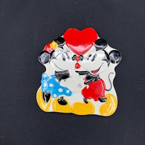 May include: A ceramic wall plaque featuring a raised, glossy illustration of two cartoon mice facing each other. One mouse wears a blue polka-dot skirt and a yellow bow, while the other wears red shorts. A large, bright red heart is positioned above them.