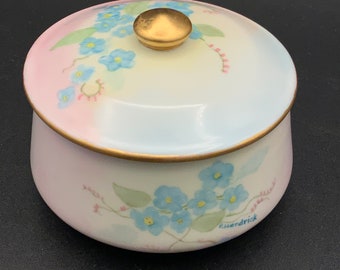 May include: A white porcelain jar with a gold lid and floral design. The jar is decorated with blue forget-me-not flowers and pink accents. The bottom of the jar is signed "Rhendrick".