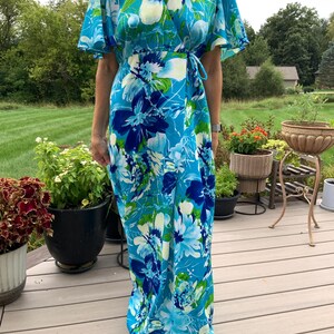 Puede incluir: Un vestido cruzado largo hasta el suelo con un vibrante estampado floral en tonos turquesa, azul real y verde lima. La prenda presenta un escote en V, mangas cortas con vuelo y un lazo lateral que asegura el panel frontal cruzado.