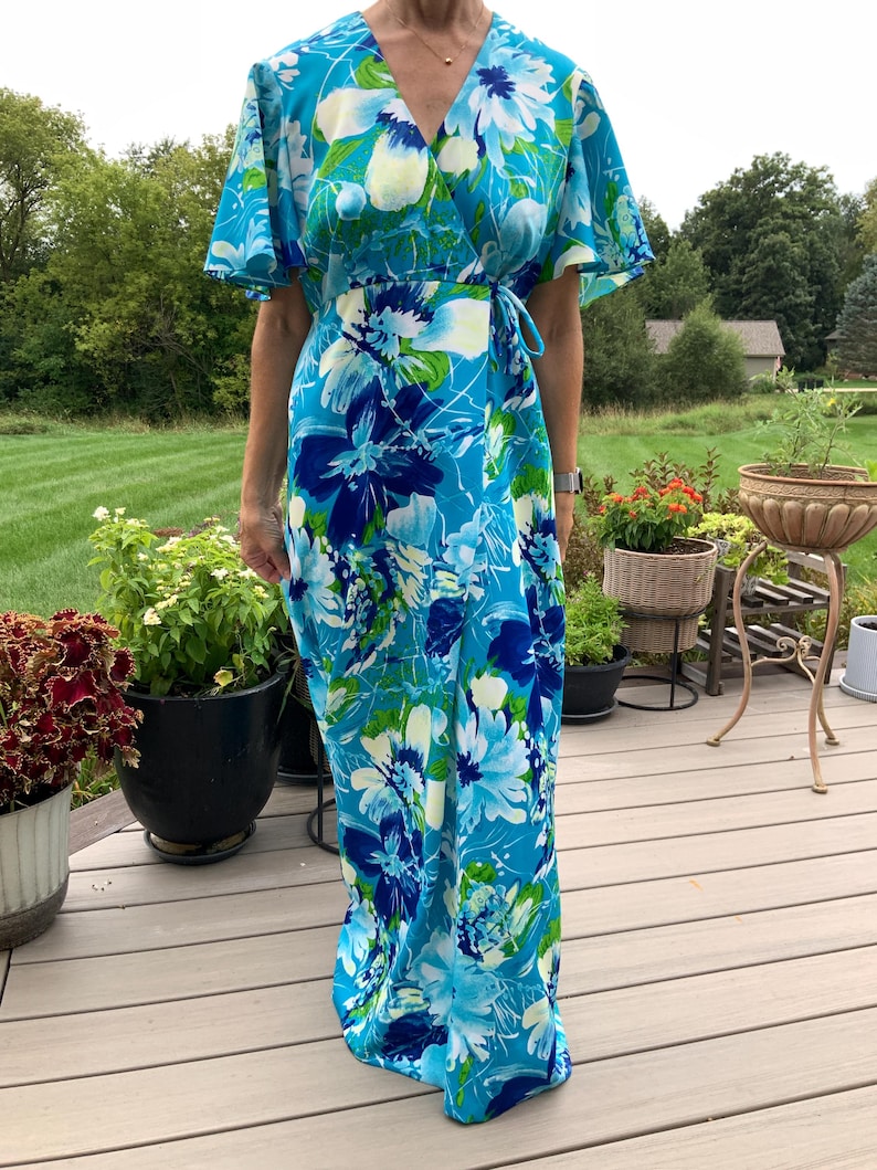Puede incluir: Un vestido cruzado largo hasta el suelo con un vibrante estampado floral en tonos turquesa, azul real y verde lima. La prenda presenta un escote en V, mangas cortas con vuelo y un lazo lateral que asegura el panel frontal cruzado.
