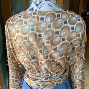 K&ouml;nnte beinhalten: Lang&auml;rmliges Oberteil mit einem Retro-Geometrie-Muster in Braun-, Beige- und Wei&szlig;t&ouml;nen. Das Oberteil ist in der Taille gerafft und wird mit einer blauen Jeans kombiniert. Der Stoff hat einen leichten Glanz.
