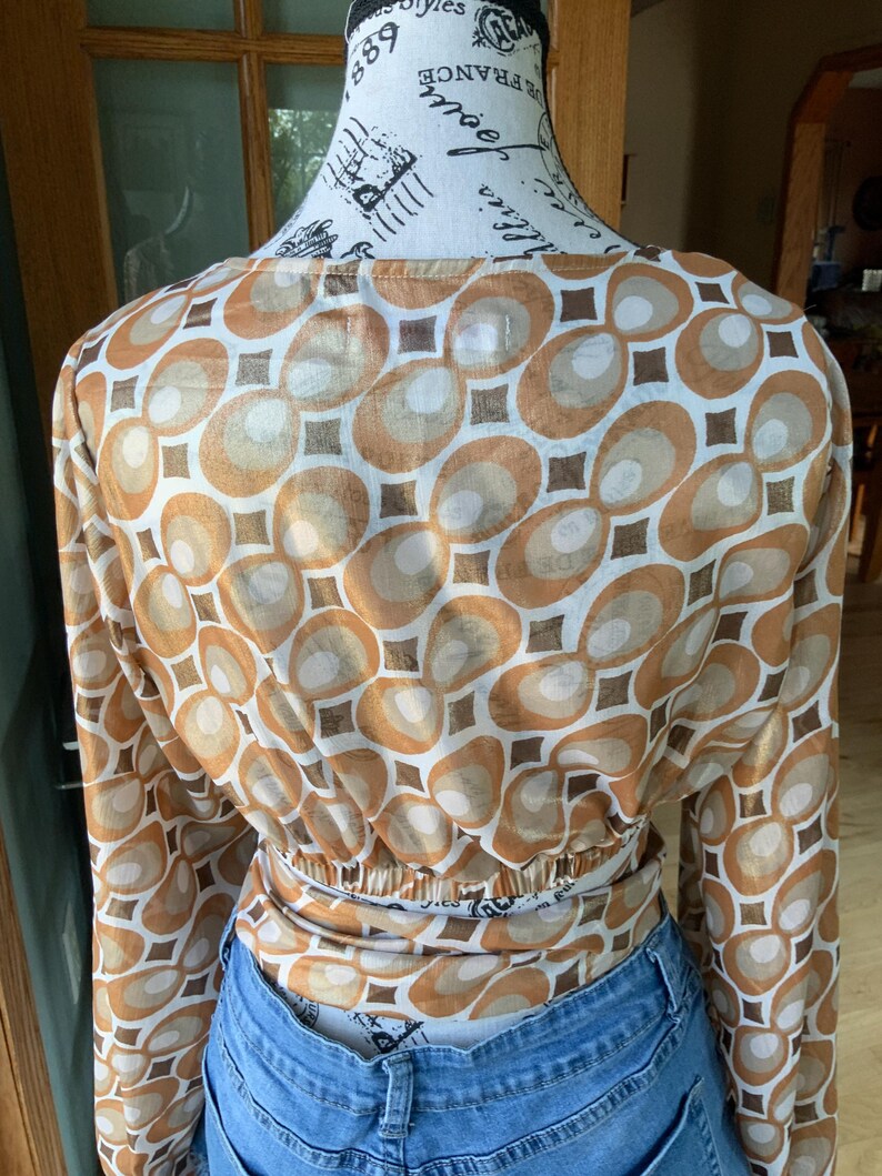 K&ouml;nnte beinhalten: Lang&auml;rmliges Oberteil mit einem Retro-Geometrie-Muster in Braun-, Beige- und Wei&szlig;t&ouml;nen. Das Oberteil ist in der Taille gerafft und wird mit einer blauen Jeans kombiniert. Der Stoff hat einen leichten Glanz.
