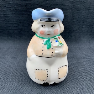 May include: A ceramic figurine of a person wearing a blue hat, tan jacket, and white pants with tan patches. The figurine is holding a small bouquet of flowers and has a green scarf.