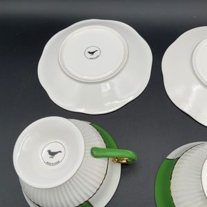 May include: Set of four white teacups and saucers with green and gold trim. The cups and saucers have a fluted design and feature a small bird logo on the bottom. The logo reads "Made in China".