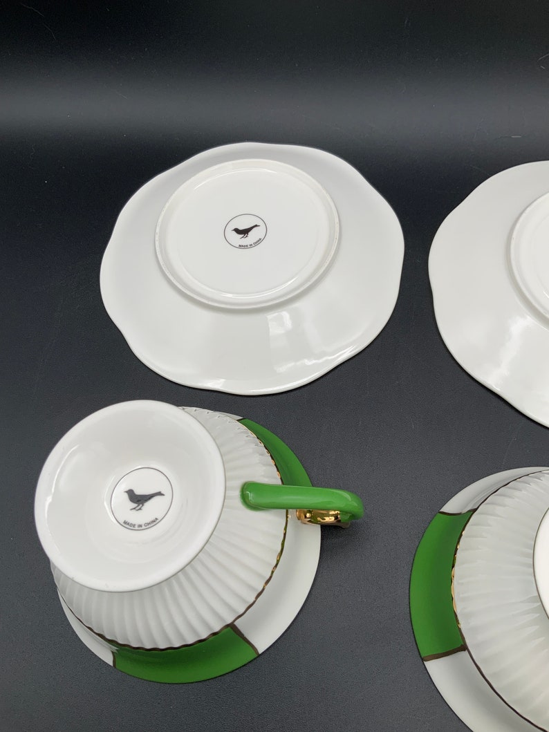 May include: Set of four white teacups and saucers with green and gold trim. The cups and saucers have a fluted design and feature a small bird logo on the bottom. The logo reads "Made in China".
