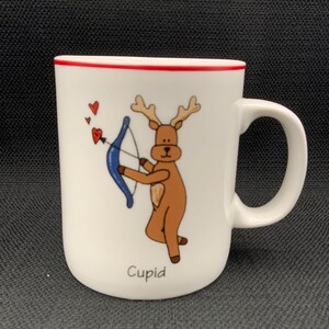 May include: White ceramic mug with a red rim, featuring a cartoon reindeer dressed as Cupid. The reindeer holds a blue bow and arrow, with three red hearts above. The word "Cupid" is printed below the reindeer.