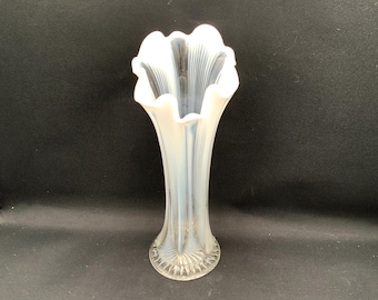 May include: A tall, slender opalescent glass vase featuring a flared, ruffled rim and vertical ribbed detailing along the body. The base is circular with a radiating pattern, and the glass transitions from a translucent clear to a milky white hue.