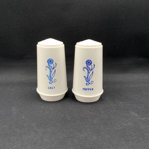 Puede incluir: Un par de saleros y pimenteros de color blanco roto con un estampado floral azul en la parte frontal. Cada recipiente tiene una forma cilíndrica cónica con una tapa perforada. La palabra SALT está impresa en azul en uno y PEPPER en el otro.