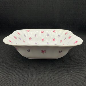 May include: A white porcelain serving bowl with a scalloped rim. The bowl is decorated with small pink and blue floral patterns. The bowl has a rectangular shape with slightly curved sides.