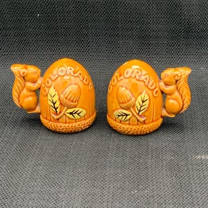 May include: A pair of ceramic salt and pepper shakers shaped like acorns with squirrels on top. The shakers are brown with yellow accents and the word "COLORADO" is written on each shaker.