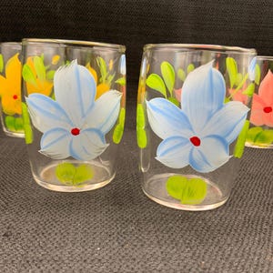 Puede incluir: Juego de cuatro vasos de vidrio transparente con diseños florales pintados a mano. Cada vaso presenta una flor de color diferente con hojas verdes y un centro rojo.