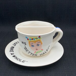 Könnte beinhalten: Ein cremefarbenes Tassen- und Untertassenset aus Keramik. Die Tasse verfügt über einen halbkreisförmigen Innenteiler und eine bemalte Illustration einer Person mit blonden Haaren. Der Text lautet HIS BETTER HALF und THAT MAKE A WHOLE.