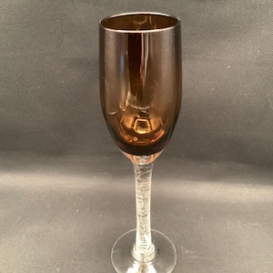 May include: A tall, slender glass with a deep amber-colored bowl and a clear, textured stem featuring a distinct crackled glass pattern. The glass has a smooth, rounded rim and a wide, circular base, standing upright with a refined, elegant profile.