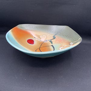 May include: A decorative ceramic bowl with a unique, abstract design. The bowl features a turquoise rim and a center with orange, tan, and green hues. A red dot and black and blue lines add to the artistic flair.