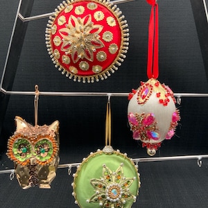Puede incluir: Cuatro adornos navideños hechos a mano, incluyendo un adorno redondo rojo y dorado, un adorno redondo blanco y rosa, un adorno redondo verde y dorado y un adorno de búho marrón y dorado.