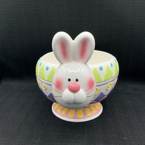 Può includere: Una ciotola a forma di coniglietto pasquale in ceramica bianca con un volto di coniglio sul davanti. La ciotola presenta un design a uovo colorato con accenti verdi, gialli e viola. Il coniglietto ha guance rosa e naso rosa.