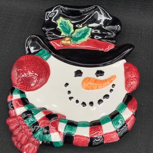 Puede incluir: Un plato de cerámica con forma de muñeco de nieve con un sombrero negro, una bufanda roja y acebo verde. El muñeco de nieve tiene una nariz de zanahoria y ojos negros.