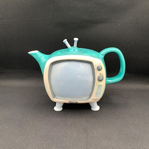 May include: A teal and cream ceramic teapot shaped like a vintage television set. The teapot has a blue screen, a handle, and two small antenna. The base has four small feet.