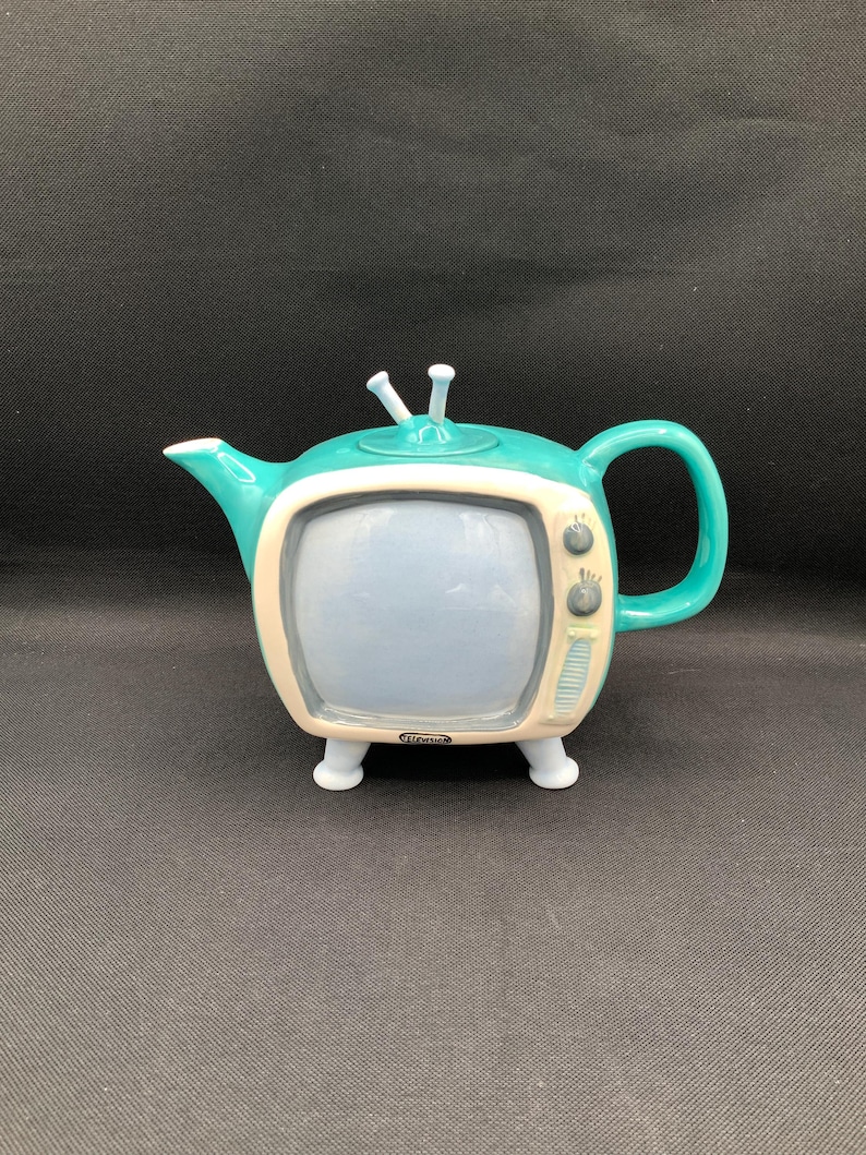 May include: A teal and cream ceramic teapot shaped like a vintage television set. The teapot has a blue screen, a handle, and two small antenna. The base has four small feet.