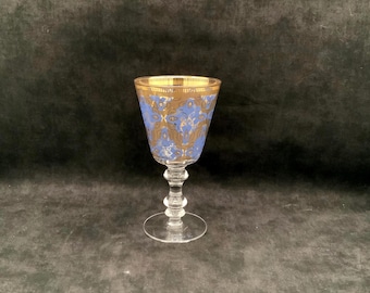 May include: A clear glass goblet featuring a blue and gold patterned design on the bowl. The rim is accented with a gold band. The stem has a multi-tiered, rounded shape leading down to a circular, clear glass base for stability.