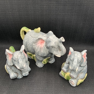 May include: A ceramic tea set featuring three elephant figurines. The teapot is a larger elephant with a green leaf design. The two smaller elephants are creamers or sugar bowls. The elephants are gray with pink ears and green leaves.