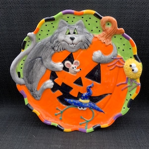 Puede incluir: Un plato de cerámica con temática de Halloween con un gato gris sonriente sobre una calabaza naranja. El plato tiene un borde verde con lunares y presenta un ratón, una araña y un sombrero de bruja. Una decoración festiva.