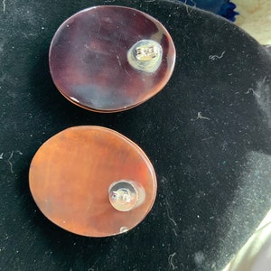 May include: Two oval-shaped, translucent earrings. One is a deep burgundy color, and the other is a lighter, reddish-brown. Each earring has a silver-colored post on the back. The earrings are displayed on a black surface.