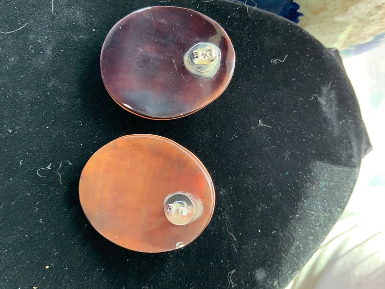 May include: Two oval-shaped, translucent earrings. One is a deep burgundy color, and the other is a lighter, reddish-brown. Each earring has a silver-colored post on the back. The earrings are displayed on a black surface.