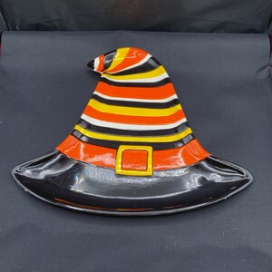 Puede incluir: Un plato de servicio de cerámica con forma de sombrero de bruja que presenta rayas horizontales en naranja, amarillo, blanco y negro. La base del sombrero incluye una banda naranja ancha con una hebilla cuadrada amarilla y un borde negro brillante.