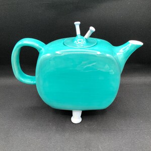 May include: A teal ceramic teapot with a rounded square shape, a curved handle, and a spout. The lid has two white decorative elements. The teapot rests on a single white foot.