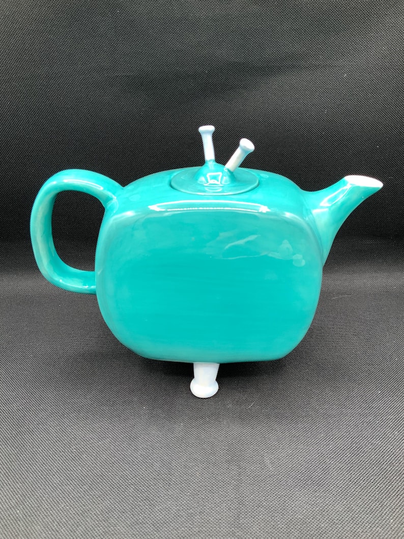 May include: A teal ceramic teapot with a rounded square shape, a curved handle, and a spout. The lid has two white decorative elements. The teapot rests on a single white foot.