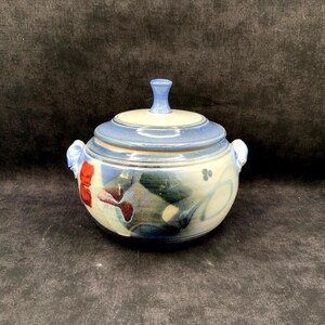 May include: A round ceramic lidded bowl featuring a cream-colored base with blue and red abstract glaze patterns. The bowl has two side handles and a matching lid with a central knob handle, showcasing a smooth, glossy finish throughout the piece.