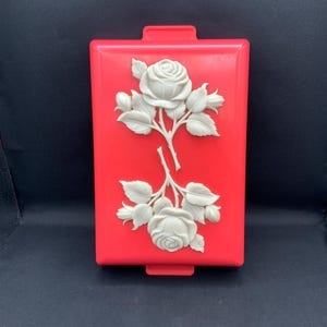 Könnte beinhalten: Ein rechteckiger roter Kunststoffbehälter mit zwei erhabenen, cremeweißen Blumenreliefs. Jedes Motiv zeigt eine blühende Rose, umgeben von Blättern und Knospen. Der Behälter verfügt über kleine Laschen an den oberen und unteren Kanten.