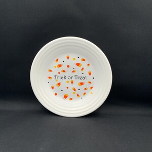 May include: A white ceramic bowl with a ribbed rim featuring a pattern of orange, yellow, and white candy corn shapes, small orange dots, and black dots. The text "Trick or Treat" is printed in black in the center of the bowl interior.