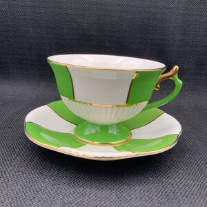 May include: A white teacup and saucer with green and gold stripes. The cup has a gold rim and a handle. The saucer has a scalloped edge.