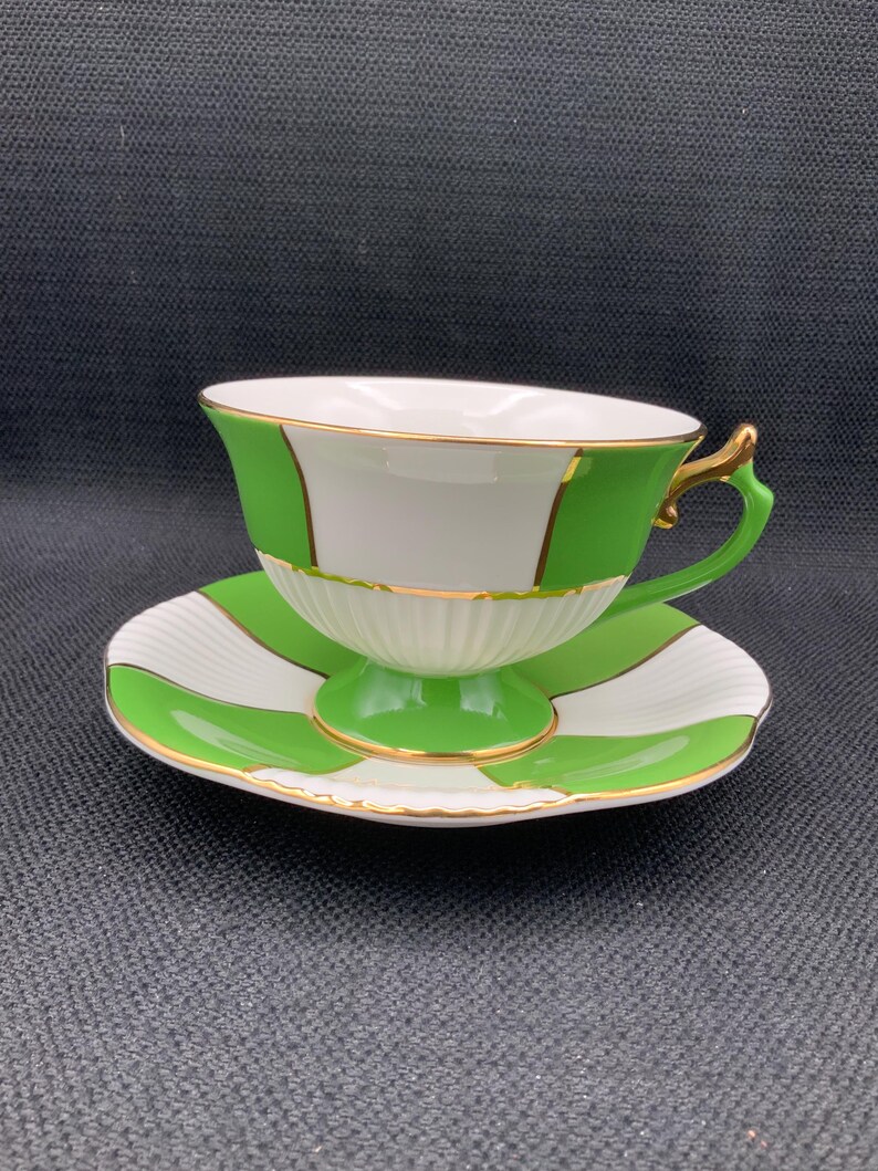 May include: A white teacup and saucer with green and gold stripes. The cup has a gold rim and a handle. The saucer has a scalloped edge.