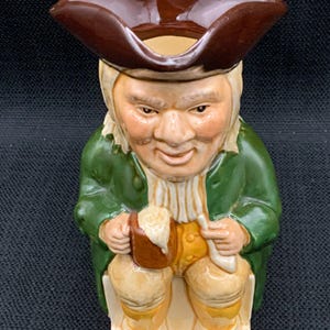 Puede incluir: Una jarra de cerámica con forma de hombre que lleva un sombrero marrón, una chaqueta verde y pantalones amarillos. Está sosteniendo una taza marrón con espuma blanca.
