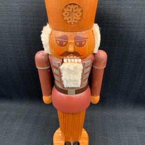 Könnte beinhalten: Eine Holz-Nussknacker-Figur in Rot- und Brauntönen. Die Figur hat einen hohen Hut mit Sternmuster, einen weißen Bart und ein bemaltes Gesicht. Sie steht auf einem Holzsockel, eine klassische Weihnachtsdekoration.