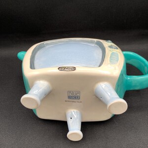 May include: A ceramic teapot shaped like a vintage television set. The teapot is primarily white with teal accents, including the handle and details. The word "TELEVISION" is printed on the front. The bottom of the teapot has four light blue legs.