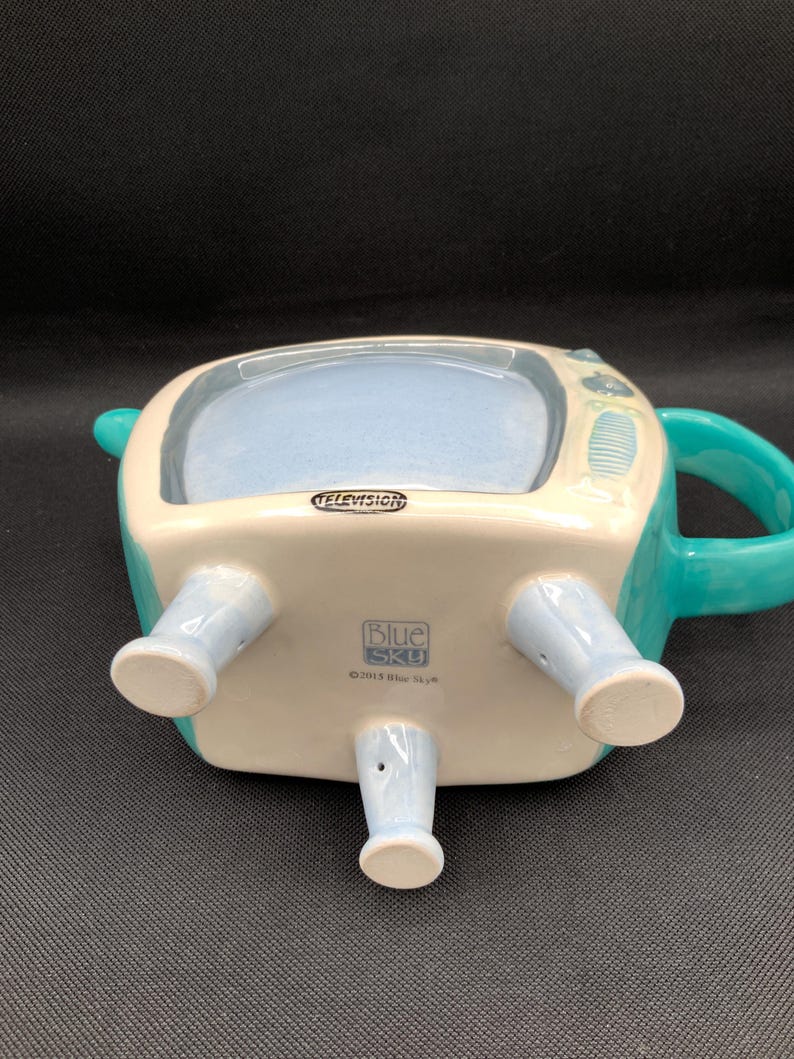 May include: A ceramic teapot shaped like a vintage television set. The teapot is primarily white with teal accents, including the handle and details. The word "TELEVISION" is printed on the front. The bottom of the teapot has four light blue legs.