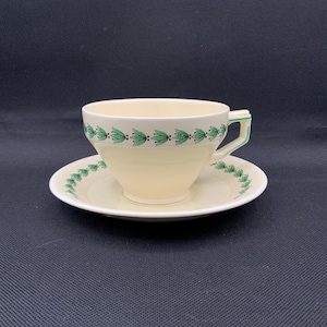 Puede incluir: Juego de taza y platillo de té color crema con un patrón de hojas verdes. La taza tiene forma redondeada con asa, y el platillo es circular. El diseño de hojas verdes se repite alrededor del borde de la taza y el platillo. El asa tiene un acento verde.