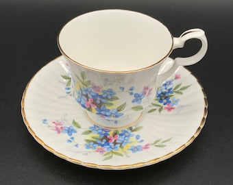 May include: A white teacup and saucer set with a floral pattern featuring blue, pink, and yellow flowers. The cup and saucer have a gold trim.