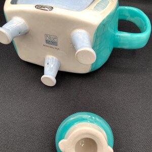 May include: A ceramic mug shaped like a vintage television set, with a light blue interior and a teal exterior. The mug has a handle and four light blue legs. The word "TELEVISION" is printed on the top. The lid is teal and white.