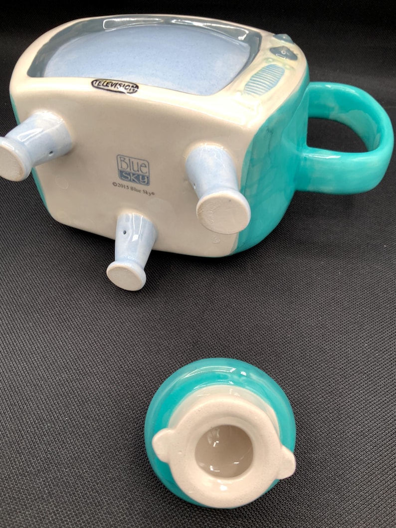 May include: A ceramic mug shaped like a vintage television set, with a light blue interior and a teal exterior. The mug has a handle and four light blue legs. The word "TELEVISION" is printed on the top. The lid is teal and white.