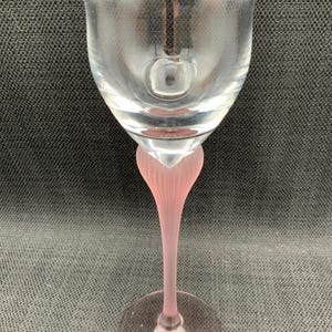 Pode incluir: Uma taça de vinho de vidro transparente com haste e base rosa. A haste tem um design canelado e a base é redonda. A taça tem formato de tulipa. O vidro é transparente.