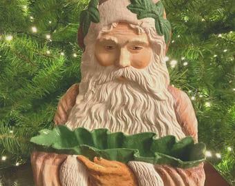 Op de afbeelding: Een keramieken Santa Claus beeldje met een lange witte baard, een bruine hoed en een groene bladkroon. Het beeldje houdt een met groene bladeren gevoerde mand vast. De achtergrond is een kerstboom.