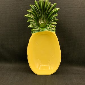May include: A ceramic pineapple-shaped dish. It has a bright yellow body and a green leafy top. The dish is designed to resemble a pineapple fruit, with a textured surface and a shallow bowl shape.